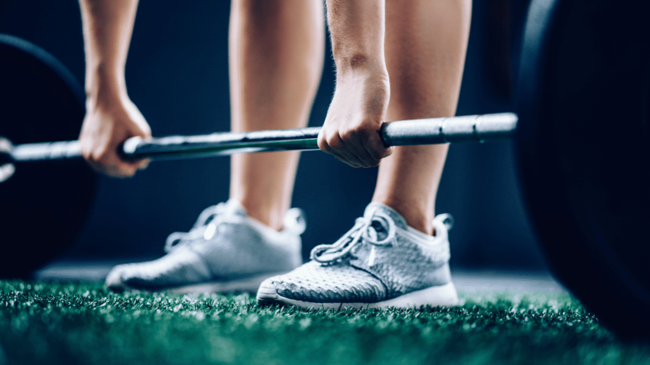 The legs of a woman engaged in functional strength training, lifting a barbell.