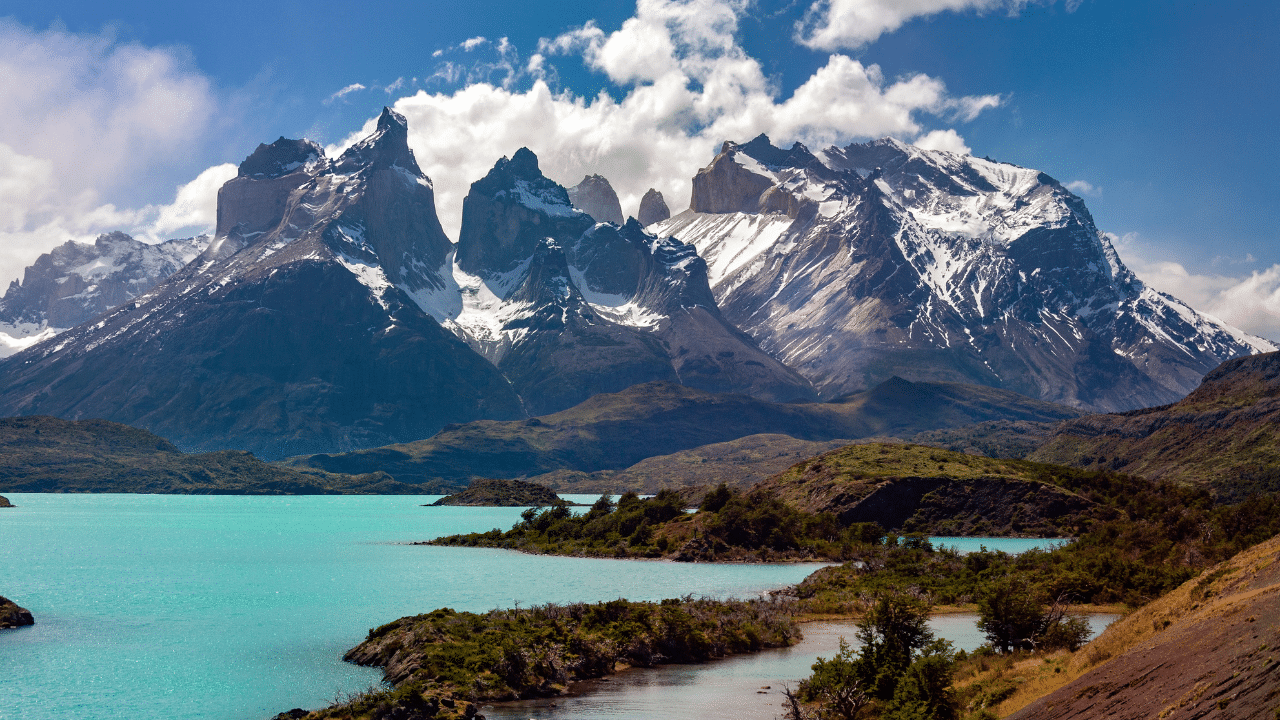 Snow-capped mountain range with jagged peaks overlooks a turquoise lake and green valley under a partly cloudy sky, the perfect backdrop for an International Marathon event in Patagonia.