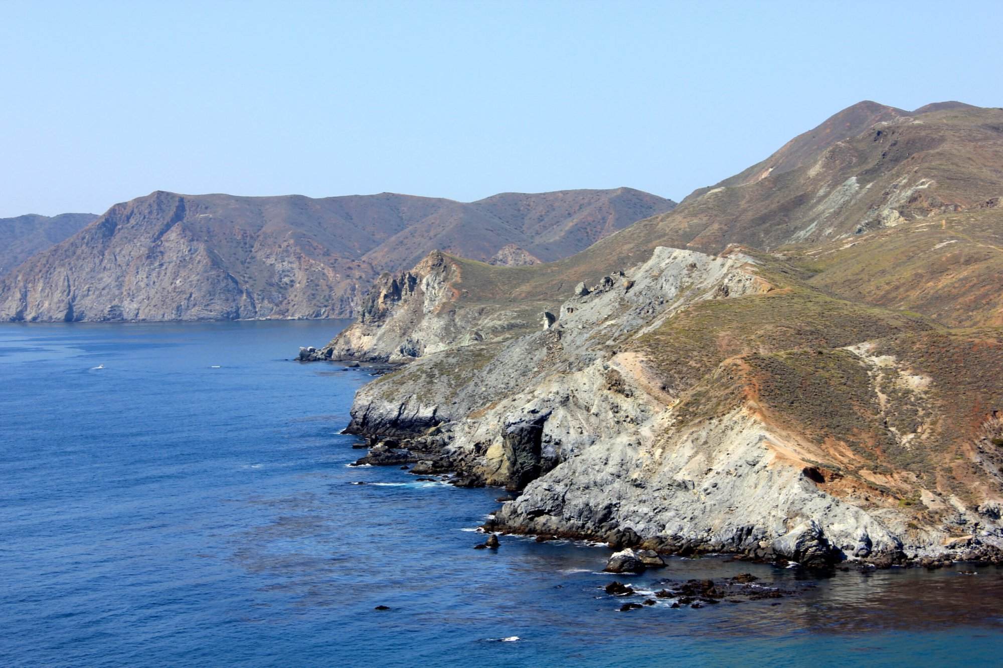 Rocky coastal cliffs with rugged terrain and sparse vegetation border a calm blue sea under a clear sky, perfect for daring adventurers tackling the Avalon Benefit 50 Mile Run.