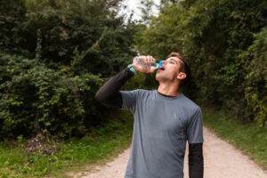 A man in a gray shirt drinks from a water bottle while standing on a forest path, surrounded by trees and foliage. As he prepares for his first ultramarathon, he follows his training guide closely during these crucial moments.