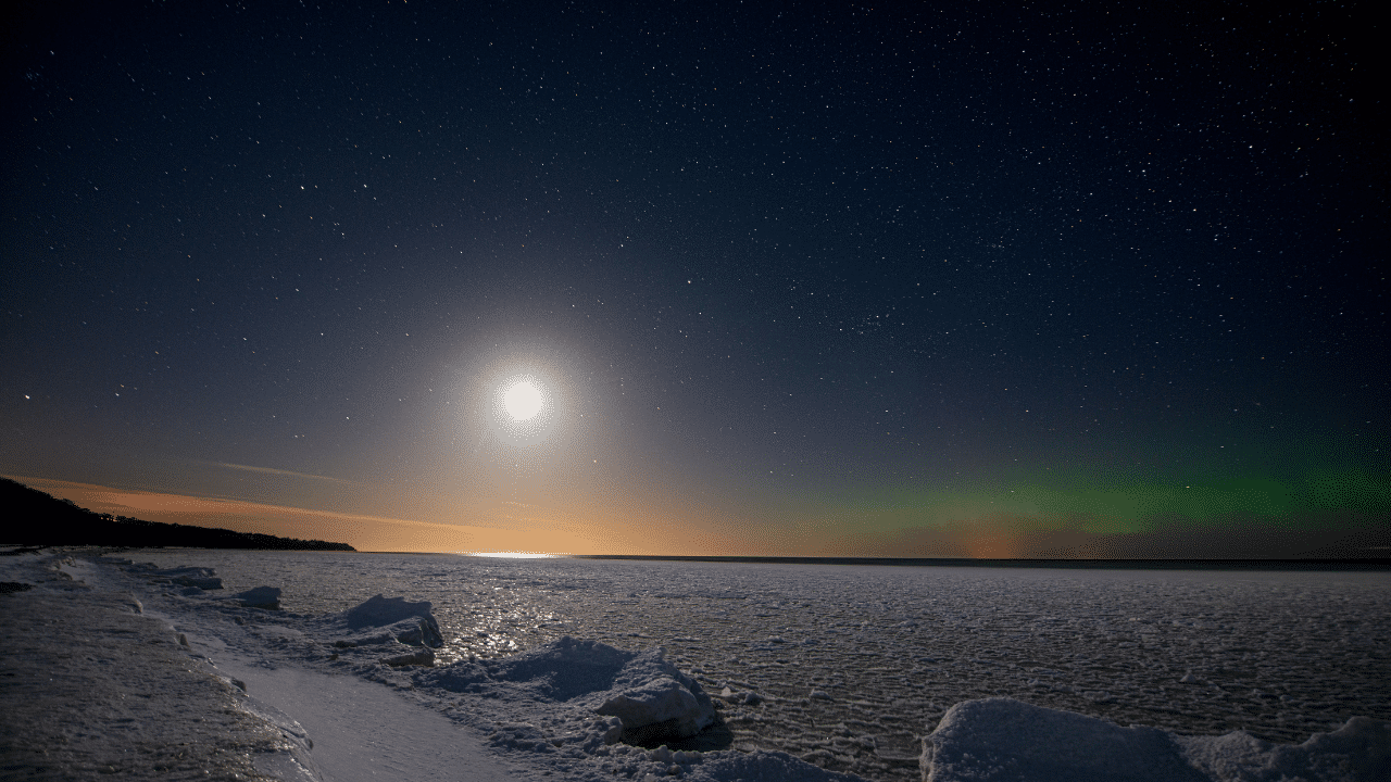 A full moon lights up a snowy coastline under a star-filled night sky with the Northern Lights visible on the horizon, creating an enchanting backdrop for the Polar Night Marathon.