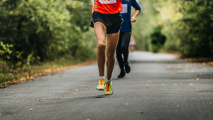 Two runners on an outdoor path, with the focus on the runner in the foreground wearing bright neon shoes and an orange shirt. The path is surrounded by dense greenery, perfect for those running their first race, from 5K to marathon.