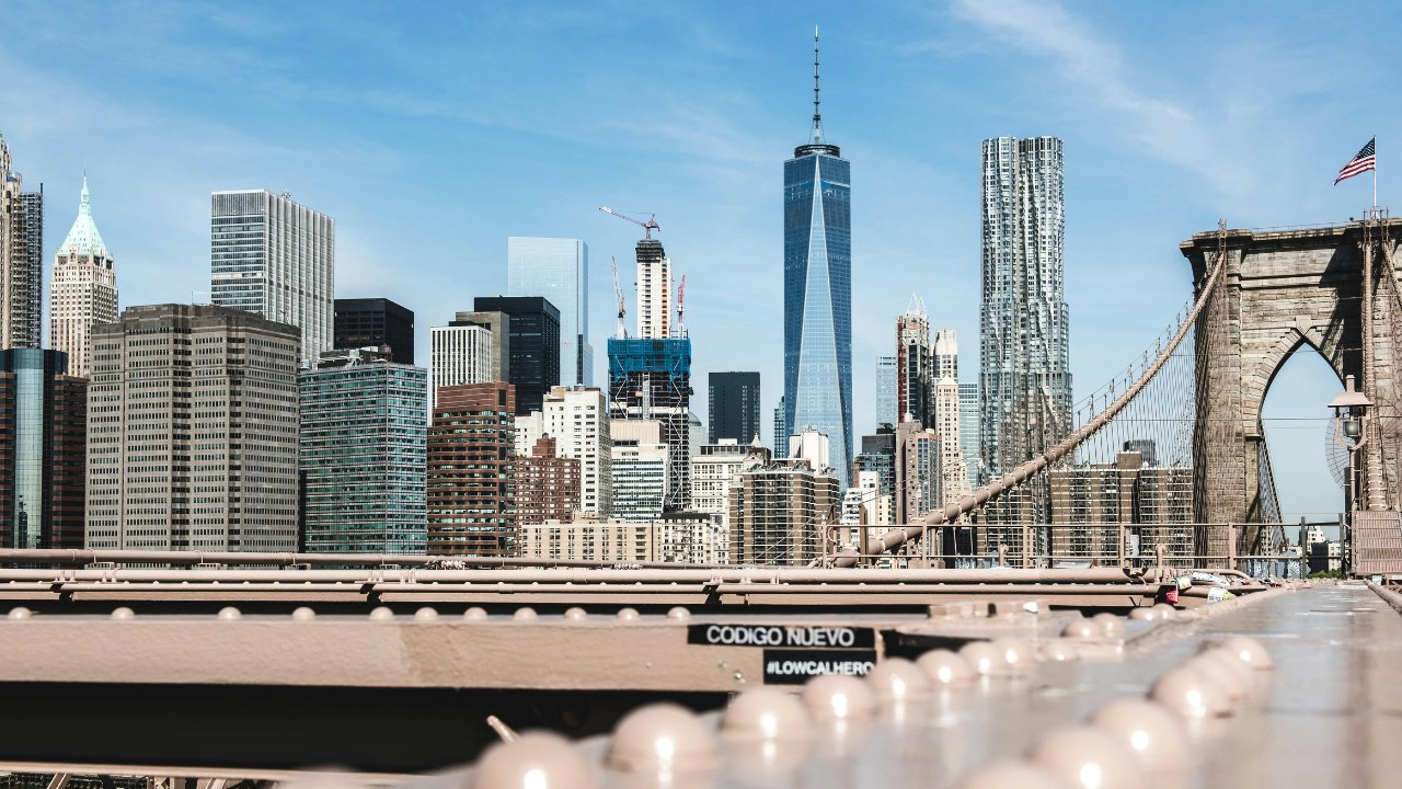 View of the New York City skyline featuring One World Trade Center and surrounding skyscrapers. The Brooklyn Bridge, often part of the route for a popular running race, is visible in the foreground under a clear blue sky.